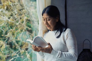 Olgun İspanyol kadın bir kitap okumaya dalmış, yemyeşil bir bahçeye bakan pencerenin yanında duruyor, huzurlu ve huzurlu bir atmosfer yaratıyor.