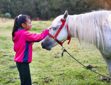 Gözlüklü ve pembe ceketli genç bir kız canlı yeşil bir alanda kırmızı halkalı güzel beyaz bir atı nazikçe evcilleştiriyor, insan ve hayvan arasında iç açıcı bir bağ kuruyor.