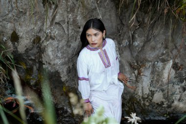 Renkli nakış işlemeli geleneksel beyaz elbiseler giyen yerli kadın kayalık bir nehirde poz veriyor. Kültürel mirası ve doğayla olan bağlantısını gösteriyor.