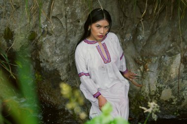 Geleneksel giysiler giyen yerli bir kadın, atalarının ve kimliğinin güzelliğini yansıtırken kayalık bir nehrin kenarında, kültürel mirası ve doğayla olan derin bağlantısını somutlaştırıyor.