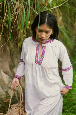 Geleneksel beyaz işlemeli yerli kadın canlı bir tropikal yağmur ormanında dolaştığı sırada kültürel miras ve doğa bağlantısını yansıtan dokunmuş bir sepet taşıyor.