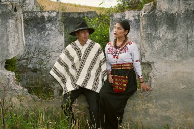 Geleneksel Andean giysileri giyen yerli kadın ve erkek harabe beton bir duvarın yanında poz veriyor, zengin kültürel miraslarını ve atalarının topraklarıyla olan bağlantılarını sergiliyorlar.