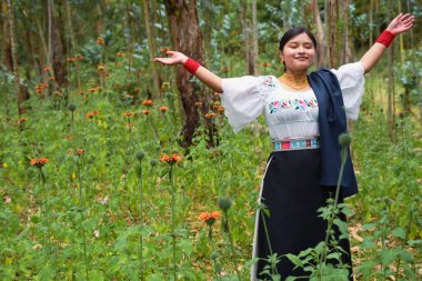 Geleneksel beyaz işlemeli bluz, siyah etek ve mavi şal giyen yerli Kolombiyalı kadın yemyeşil bir ormanda turuncu kır çiçekleriyle sükuneti kucaklıyor.