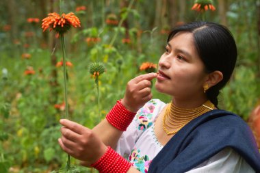Geleneksel giyinmiş yerli bir kadın ve karmaşık mücevherlerle süslenmiş, yemyeşil bir tarlanın ortasında canlı turuncu bir çiçeğin zengin kokusunu nazikçe içine çekiyor.