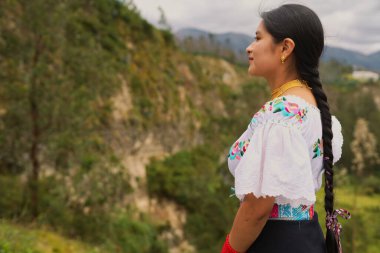 Yerli Kolombiyalı kadın, canlı, nakışlı geleneksel giysiler ve altın takılar ile süslenmiş, profilde duruyor, serin yeşil dağlık manzaraya bakıyor