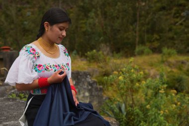 Geleneksel Andean giysileri ve altın mücevherleri giyen genç otavalo kadın elinde bir parça koyu mavi kumaş tutuyor ve ekvatorun yüksek kesimlerinde doğal bir ortamda poz veriyor.