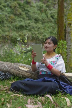 Geleneksel giysiler giyen yerli bir kadın, doğal ortamında dijital tablet kullanarak düşmüş bir kütüğün yanında çimlerin üzerinde oturur ve huzurlu bir ortamda teknolojinin faydalarından keyif alır.