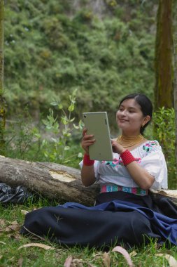 Geleneksel giysiler giyen yerli bir kadın, doğal ortamında teknolojiyi kucaklayarak, devrilmiş bir ağaç kütüğünün yakınındaki çimlerde otururken dijital tablet kullanmaktan zevk alıyor.