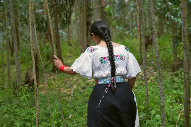 Geleneksel beyaz işlemeli bluz ve siyah etek giyen uzun siyah örgülü yerli kadın yeşil bir ormanda incecik ağaçlara dokunarak yürüyor.