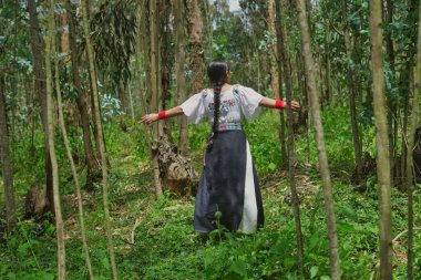 Geleneksel kıyafetler giyen yerli bir kadın gür bir ormanda kollarını açmış, doğanın sakinliğini kucaklıyor ve kültürel mirasını kutluyor.