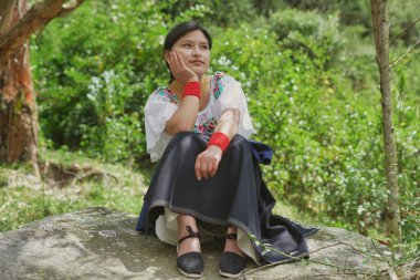 Geleneksel Andean giysileri giyen genç bir kadın huzurlu bir doğal ortamda bir kayanın üzerinde oturuyor, çevrenin güzelliğini düşünüyor ve bir anlık sakinliğin tadını çıkarıyor.