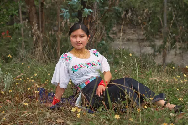 Geleneksel Andean giysileri ve altın mücevherler giyen genç yerli bir kadın sarı çiçeklerle dolu bir tarlada oturuyor ve bir portre için poz veriyor.