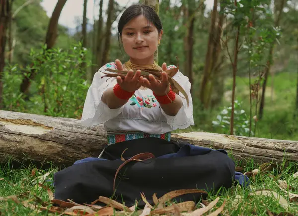 Geleneksel kıyafetlerle süslenmiş yerli bir kadın, gür bir ormanda huzur içinde oturuyor, nazikçe kuru yaprakları tutuyor, doğayla ve kültürel mirasıyla derin bir bağ kuruyor.