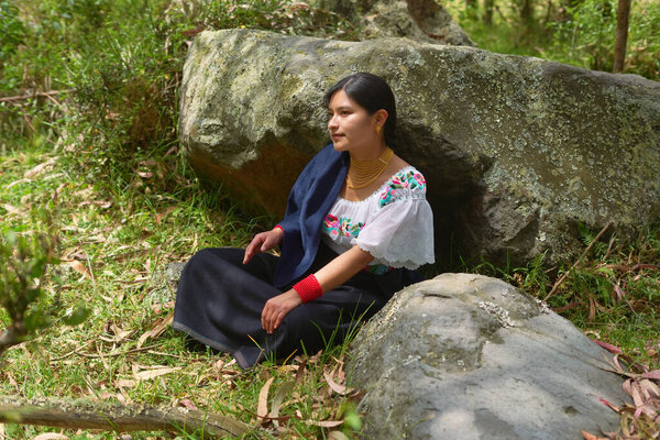 Young indigenous woman wearing traditional andean clothing sitting between rocks in a peaceful forest, showcasing her cultural heritage and connection to nature