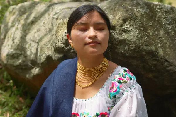Kapalı gözlü genç bir kadın geleneksel işlemeli giysiler ve altın mücevherler giyiyor arkasında büyük bir taşla doğada meditasyon yapıyor.