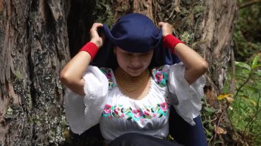 Geleneksel beyaz gömlekli ve mavi başörtülü yerli bir kadın, yaz mevsiminde bereketli orman yeşillikleriyle çevrili ağaç gövdesinin yanında dururken kültürel kıyafetlerini incelikle düzeltiyor.