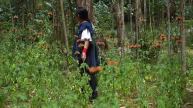 Geleneksel giysiler giymiş yerli bir kadın yeşil ormanda yürüyor, canlı turuncu çiçekleri yüksek bitkilerden hasat ediyor, doğal çevreyle derin bir bağ kuruyor.