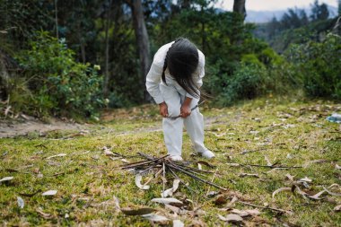 Beyaz elbiseli bir çocuk elleriyle küçük dallar kırıyor, ağaçlarla çevrili bir çayırda şenlik ateşi yakıyor, açık hava macerasının ruhunu kucaklıyor.