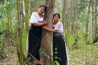 Yerli anne ve kız, geleneksel giysiler içinde, canlı bir ormanda bir ağacı kucaklayarak, onların doğa ve kültür mirasıyla derin bağlantılarını yansıtıyor.