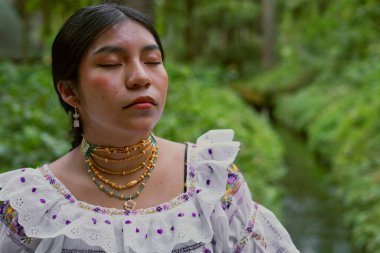 Geleneksel kıyafet ve mücevherlerle süslenmiş yerli bir kadın, sakin doğal bir ortamda gözleri kapalı duruyor, çevrenin sakinliğini kucaklıyor.