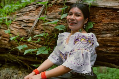 Canlı geleneksel kıyafetlerle süslenmiş yerli bir kadın gür bir ormandaki büyük bir ağaç gövdesine sessizce oturur. Gülüşü sıcaklık ve kültürel gurur yayar.