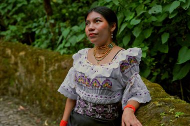 Renkli geleneksel huipil ve mücevher giyen genç bir Guatemalalı kadın canlı yeşil bir ormanda sakin poz vererek Guatemala 'nın zengin kültürel mirasını sergiliyor.