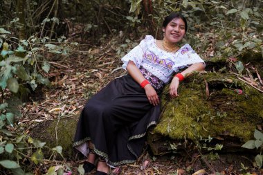Geleneksel And dekanı kıyafetleri giyen genç bir kadın gür bir ormandaki yosunlu bir kütükte dinlenirken gülümsüyor ve kültürel mirası ve doğayla bağlantısını gösteriyor.
