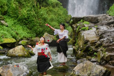 Geleneksel kıyafetler giyen iki yerli kadın bir nehirde duruyorlar ve havaya çiçek yaprakları atıyorlar, bir şelalenin önünde doğayı kutluyorlar.