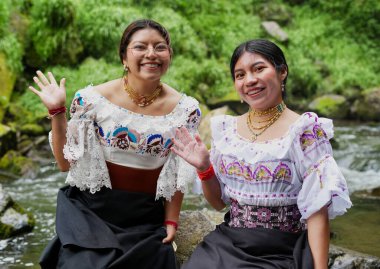 Geleneksel Guatemala kıyafetleri giyen iki neşeli yerli kadın nehir kenarında oturuyor, el sallıyor ve gülümsüyor, canlı kültürlerini ve mirasını sergiliyorlar.
