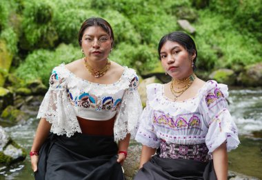 Geleneksel Guatemala kıyafetleri giyen iki yerli kadın nehir kenarında poz veriyor, canlı ve karmaşık giysilerini sergiliyorlar, zengin kültürel miraslarını temsil ediyorlar.