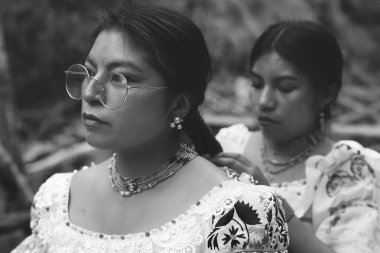 Geleneksel kıyafetler giymiş ve karmaşık mücevherlerle süslenmiş iki yerli kadın, biri diğerinin saçını ustalıkla örüyor, zengin kültürel miraslarını ve derin gururlarını kutluyorlar.