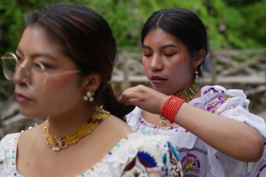 Geleneksel giysiler ve mücevherler giyen yerli bir kadın, başka bir yerli kadının saçını örüyor. Aynı zamanda geleneksel kıyafetler ve mücevherler takıyor.