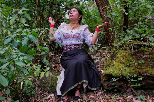 Geleneksel giysiler giyen yerli kadın gür bir ormanda yosunlu bir kayanın üzerinde oturuyor, gözleri kapalı ve elleri havada, canlı yeşil yapraklarla çevrili bir şekilde meditasyon yapıyor.