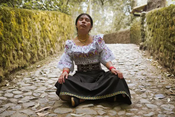 Genç yerli bir kadın, gözleri kapalı, kaldırım taşı bir yolda meditasyon yapıyor ve doğal ortamında kültürel mirasını kucaklıyor.