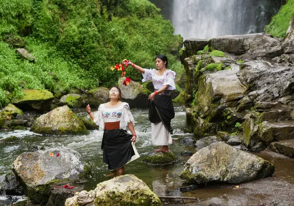 Geleneksel giyinmiş iki yerli kadın, kültürel miraslarını ve doğayla olan bağlantılarını kutlamak için şelalenin dibindeki bir nehirde renkli çiçek yapraklarıyla bir ayin yapıyorlar.