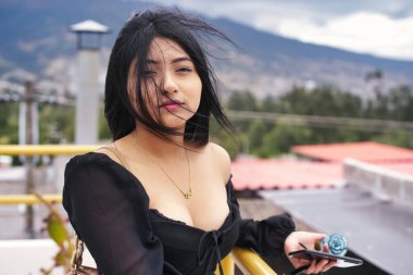 Akıllı telefonu ve arka planda manzaralı şehir manzarasıyla balkonda poz veren genç nüfuzlu kişi, moda tarzını sergiliyor ve kentsel manzaranın tadını çıkarıyor.