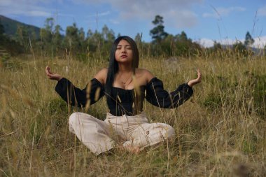 Genç bir kadın, huzurlu bir çayırda oturur, gözleri kapalı meditasyon yapar ve elleri, doğanın sükunetiyle çevrili, sakinleştirici bir Mudra 'ya yerleştirilir.