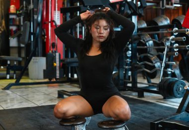 Genç atletik kadın, ağırlıklar üzerinde otururken saçını bağlıyor. Yoğun bir spor egzersizinden sonra dinleniyor. Bağlılığını ve spor yolculuğuna odaklanıyor.