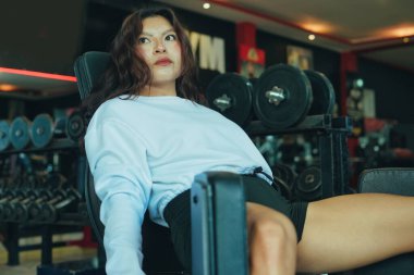 Genç bir kadın bacak egzersizi yaparken mola veriyor, makinenin üzerinde oturuyor ve modern bir spor salonunda halterlerle çevrili, spor salonuna ve sağlıklı bir yaşam tarzına olan bağlılığını gösteriyor.