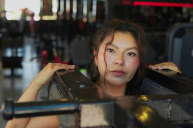 Genç bir kadın spor salonunda antreman yaparken halter makinesine yaslanıyor ve bakışlarında egzersiz ekipmanlarıyla çevrili kararlılığını gösteriyor.