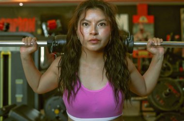 Omuzlarında ağırlıklarla çömelme hareketleri yapan genç bir kadın, kendini spora adamış bir sporcunun ruhunu somutlaştırarak, spor salonundaki forma olan bağlılığını ve gücünü gösteriyor.