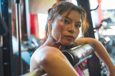 Azimli Asyalı kadın çömelme egzersizi sırasında kısa bir mola veriyor, spor ve güç eğitimine bağlılığı vurguluyor, aktif yaşam tarzında odaklanmayı ve motivasyonu somutlaştırıyor.