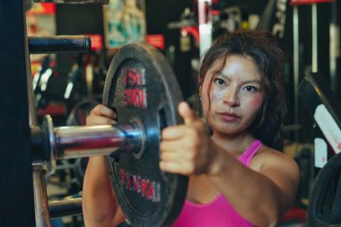 Genç bayan sporcu halterlere ağırlık ekliyor. Modern bir spor salonunda egzersiz yapmaya hazırlanıyor. Odaklanma ve kararlılık gösteriyor.