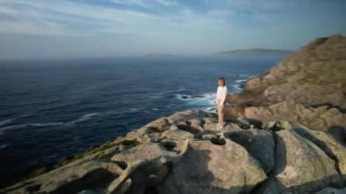 Female Tourist Standing On Rocks In Sunlight On The Coast Of Malpica In A Corua, Spain. FPV drone orbit