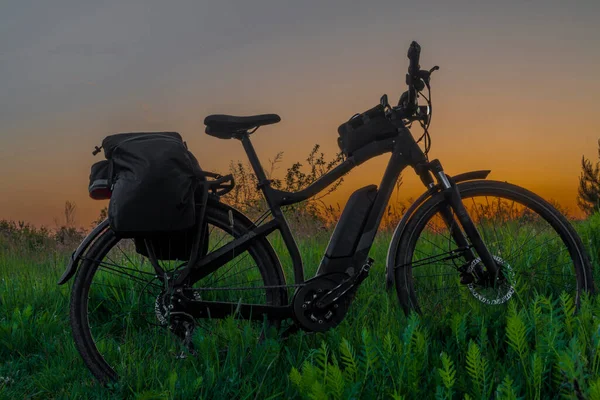 Electric bike with black bag on overgrown meadow with after sunset orange sky