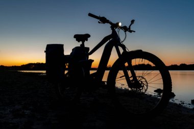 Silhouette of electric bicycle on beach of pond Bezdrev after color sunset clipart
