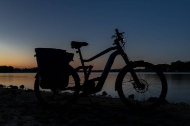 Silhouette of electric bicycle on beach of pond Bezdrev after color sunset clipart