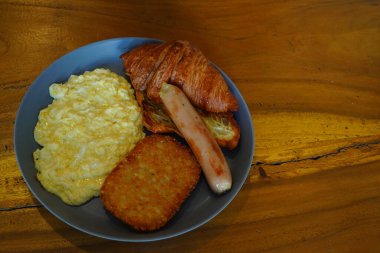 Mücver, kruvasan, sosis ve çırpılmış yumurtadan oluşan kahvaltı, ahşap bir masaya yerleştirilmiş gri seramik tabaklar üzerinde servis edilir. Yemek Fotoğrafçılığı. Boşluğu kopyala