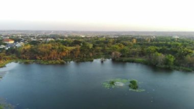 Küçük bir gölün üzerinde bir dron uçar. Dağlık arazi hatlarıyla çevrili kuru bitki örtüsüyle ve Endonezya 'nın Batı Surabaya kentindeki birkaç yüksek binanın zeminiyle. Hava görüntüleri.