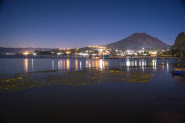 Batur Gölü, Kintamani, Bali 'deki akşam atmosferi, Batur Dağı' nın yansıması ve göl kenarındaki köyün atmosferi parlak ışıklarla aydınlatılıyor. Gece Fotoğrafçılığı.